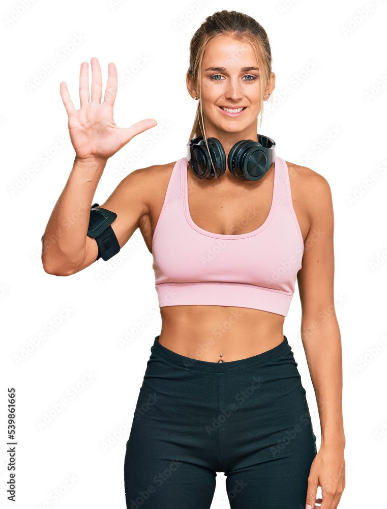 Young blonde woman wearing gym clothes and using headphones showing and pointing up with fingers number five while smiling confident and happy. Young blonde woman wearing gym clothes and using headphones showing and pointing up with fingers number five while smiling confident and happy.