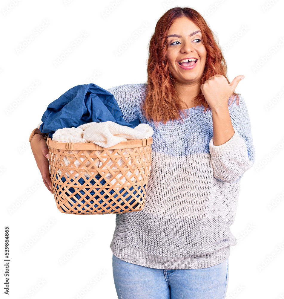 Young latin woman holding laundry basket pointing thumb up to the side smiling happy with open mouth Young latin woman holding laundry basket pointing thumb up to the side smiling happy with open mouth