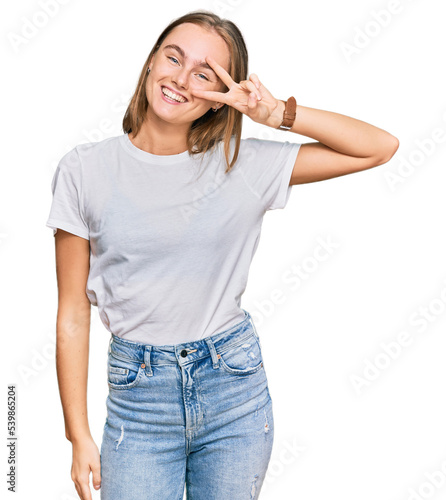 Beautiful young blonde woman wearing casual white t shirt doing peace symbol with fingers over face, smiling cheerful showing victory