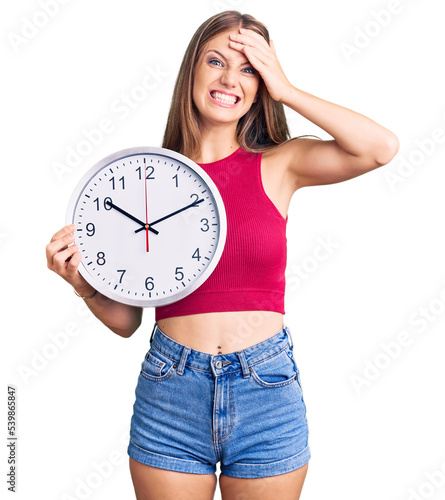 Young beautiful blonde woman holding big clock stressed and frustrated with hand on head, surprised and angry face