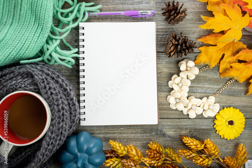 Fall flatlay, with blank spiral notebook journal, coffee mug, jewerly, scarfs and blankets. Cozy autumn scene with maple leaves on wood background