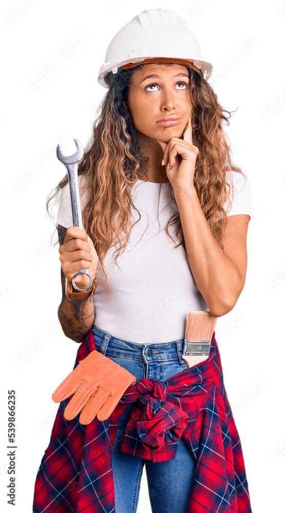 Young hispanic woman with tattoo wearing hardhat and builder clothes holding wrench serious face thinking about question with hand on chin, thoughtful about confusing idea Young hispanic woman with tattoo wearing hardhat and builder clothes holding wrench serious face thinking about question with hand on chin, thoughtful about confusing idea