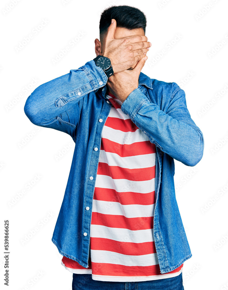 Young hispanic man with beard wearing casual denim jacket covering eyes and mouth with hands, surprised and shocked. hiding emotion Young hispanic man with beard wearing casual denim jacket covering eyes and mouth with hands, surprised and shocked. hiding emotion