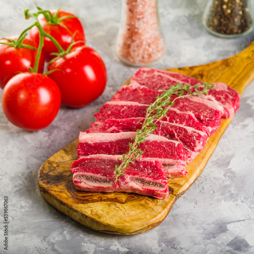On a white background, a wooden cutting board with raw meat steaks from organic meat from the farm and a sprig of fragrant rosemary, appetizing ripe tomatoes and spices in a glass jar.