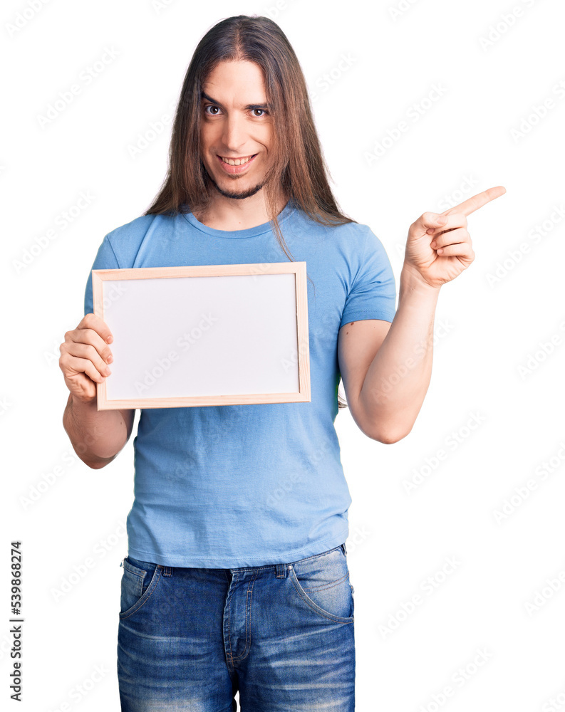 Young adult man with long hair holding empty white chalkboard smiling happy pointing with hand and finger to the side