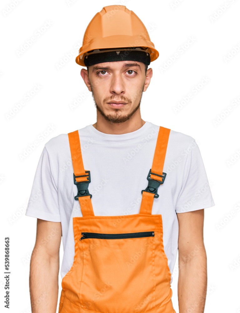 Hispanic young man wearing handyman uniform and safety hardhat with serious expression on face. simple and natural looking at the camera. Hispanic young man wearing handyman uniform and safety hardhat with serious expression on face. simple and natural looking at the camera.