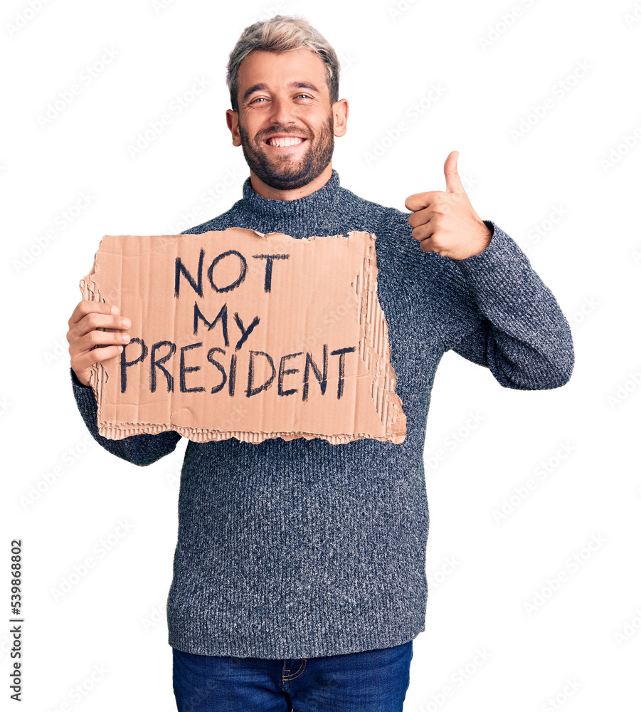 Young handsome blond man holding not my president cardboard banner smiling happy and positive, thumb up doing excellent and approval sign