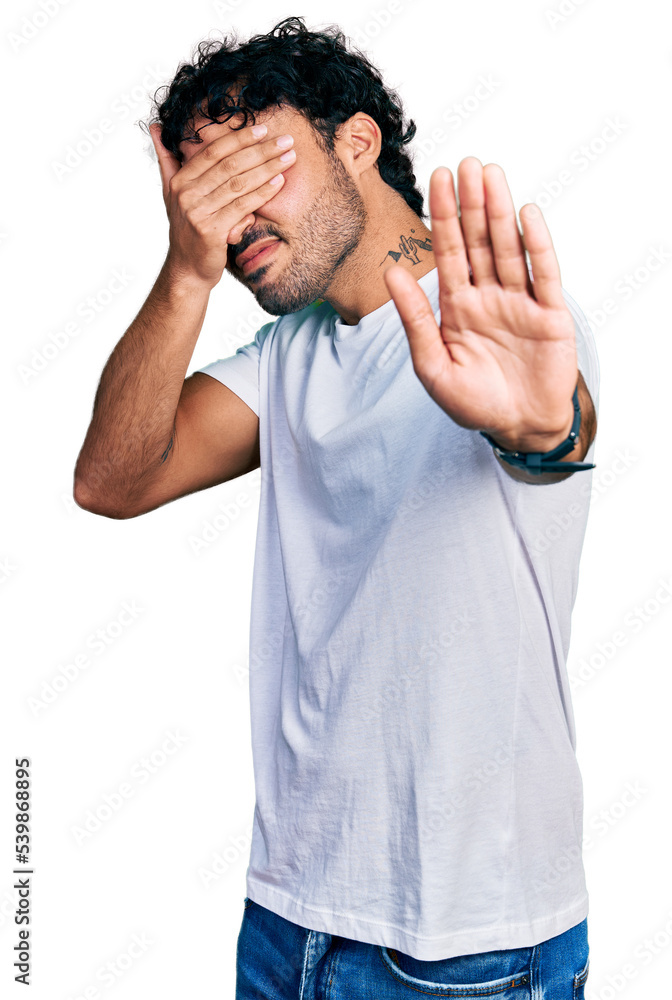 Hispanic young man with beard wearing casual white t shirt covering eyes with hands and doing stop gesture with sad and fear expression. embarrassed and negative concept. Hispanic young man with beard wearing casual white t shirt covering eyes with hands and doing stop gesture with sad and fear expression. embarrassed and negative concept.