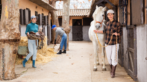 Group of female workers of stable doing daily chores in yard on autumn day, Asian woman leading white horse while girl preparing saddle for horseback ride, elderly woman stacking hay in background
