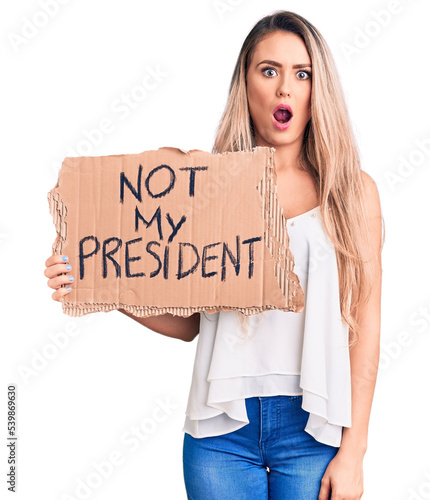 Young beautiful blonde woman holding not my president cardboard banner scared and amazed with open mouth for surprise, disbelief face