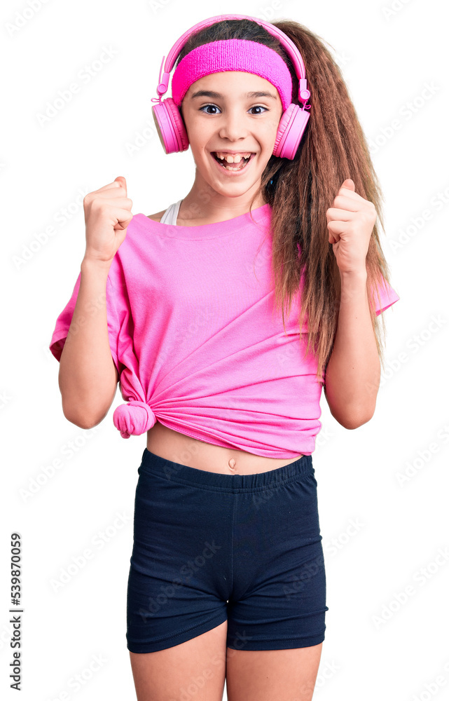 Cute hispanic child girl wearing gym clothes and using headphones celebrating surprised and amazed for success with arms raised and open eyes. winner concept. Cute hispanic child girl wearing gym clothes and using headphones celebrating surprised and amazed for success with arms raised and open eyes. winner concept.