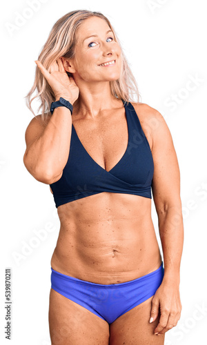 Middle age fit blonde woman wearing bikini smiling with hand over ear listening an hearing to rumor or gossip. deafness concept.