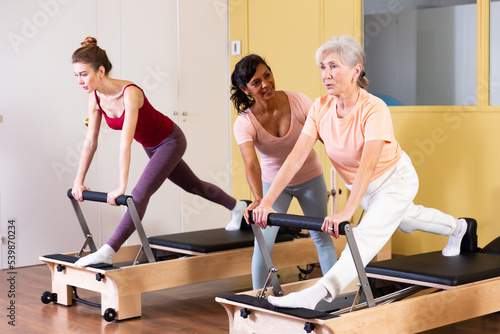 Two health conscious women of different ages perform a Pilates exercise using a reformer bed, where a female instructor ..helps them do it correctly