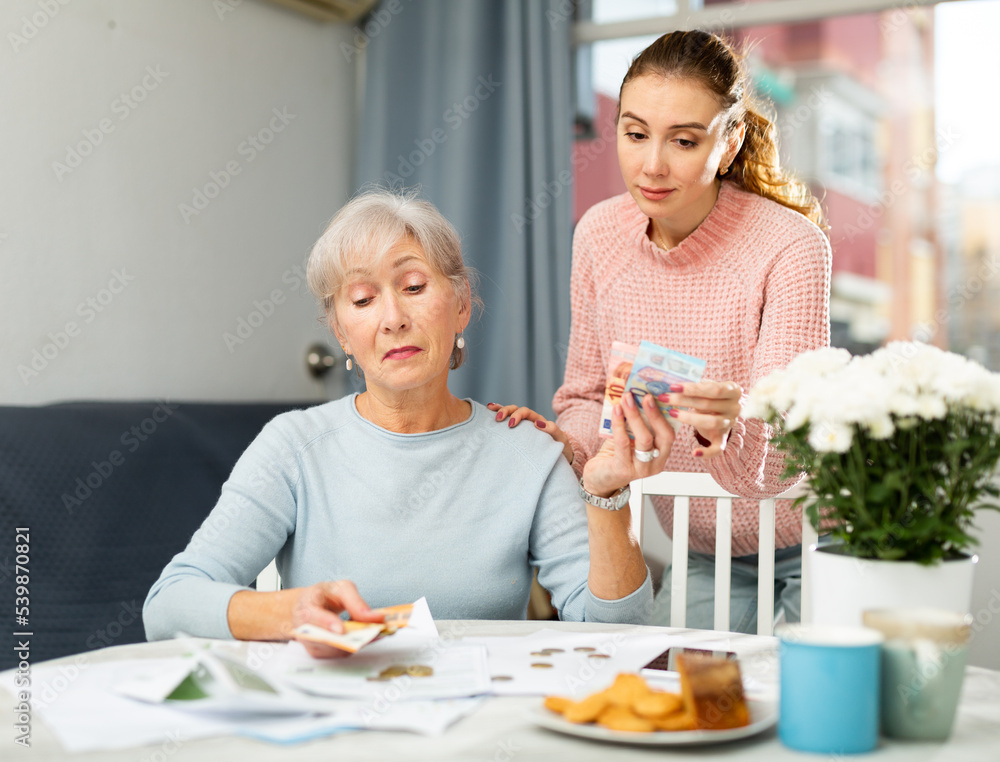Young girl asking for money from her strict elderly mother sitting at home table and calculating running costs of household chores.