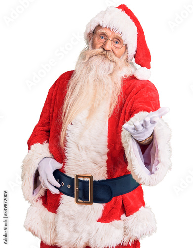 Old senior man with grey hair and long beard wearing traditional santa claus costume smiling cheerful offering palm hand giving assistance and acceptance.