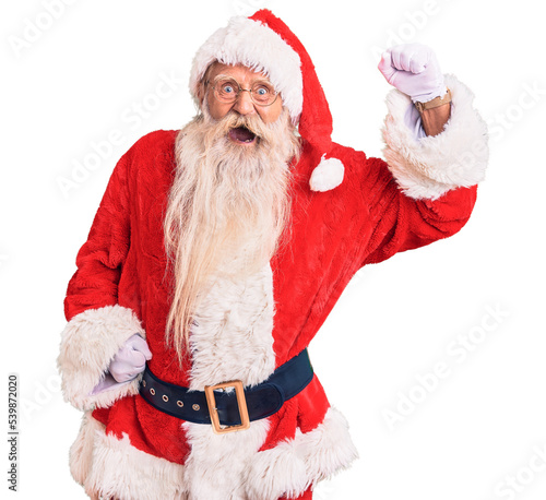 Obraz na płótnie Old senior man with grey hair and long beard wearing traditional santa claus costume angry and mad raising fist frustrated and furious while shouting with anger