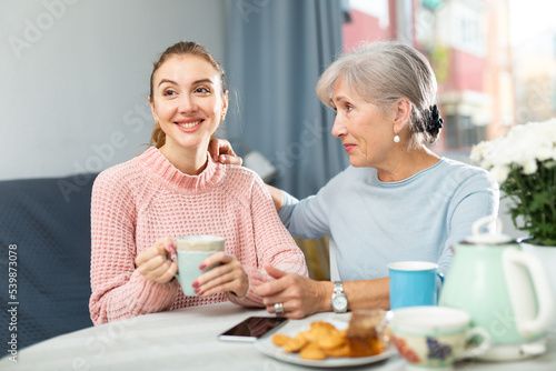 Cheerful smiling young woman enjoying conversation with elderly mother over cup of coffee with sweets at home table .