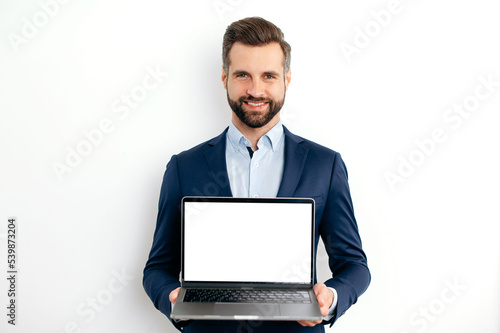 Advertisement concept. Positive caucasian bearded business man, in suit, programmer, IT specialist, seo, holds open laptop with empty mock-up screen in hand, stand on isolated white background, smiles
