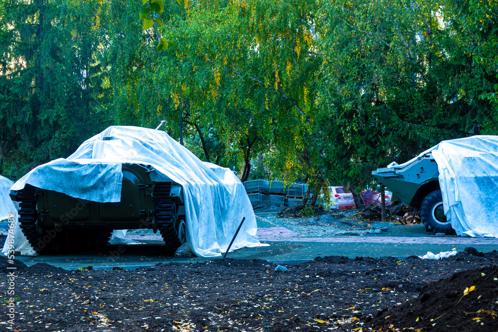 lightly armored military equipment covered with tarpaulin stands in the city park during repair work