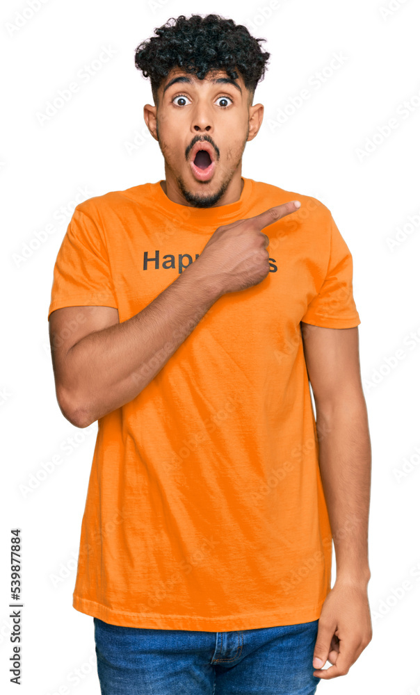 Young arab man wearing tshirt with happiness word message surprised pointing with finger to the side, open mouth amazed expression. Young arab man wearing tshirt with happiness word message surprised pointing with finger to the side, open mouth amazed expression.