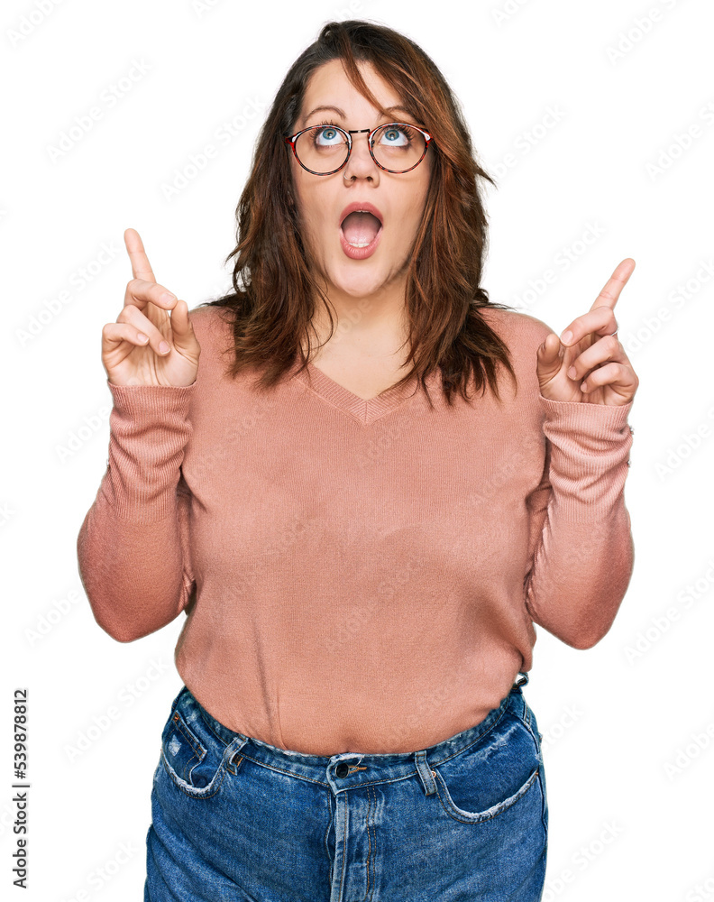 Young plus size woman wearing casual clothes and glasses amazed and surprised looking up and pointing with fingers and raised arms. Young plus size woman wearing casual clothes and glasses amazed and surprised looking up and pointing with fingers and raised arms.