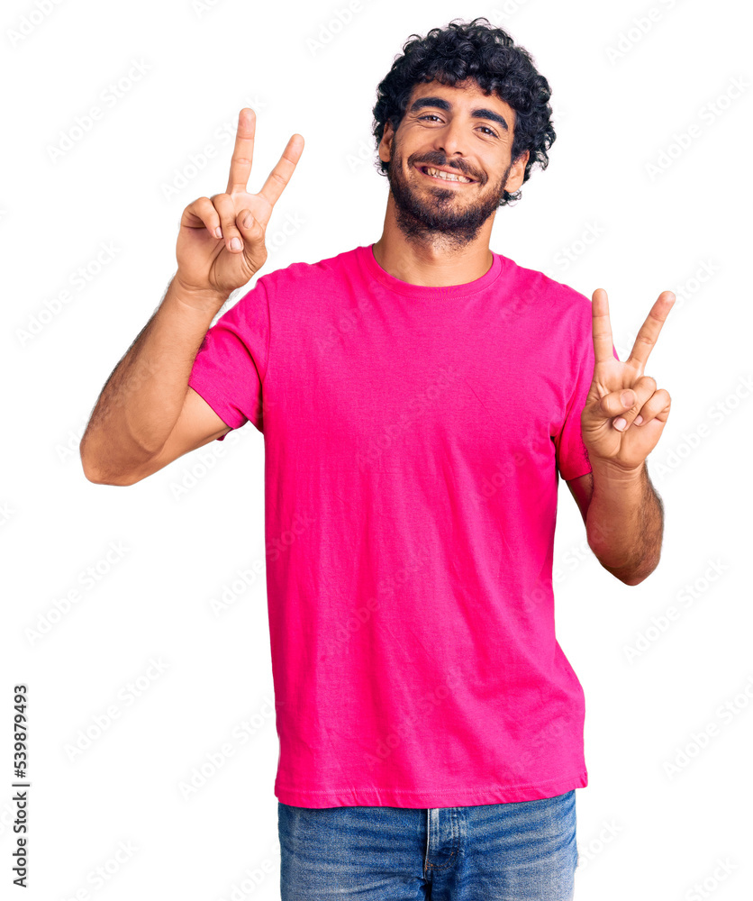 Handsome young man with curly hair and bear wearing casual pink tshirt smiling looking to the camera showing fingers doing victory sign. number two. Handsome young man with curly hair and bear wearing casual pink tshirt smiling looking to the camera showing fingers doing victory sign. number two.