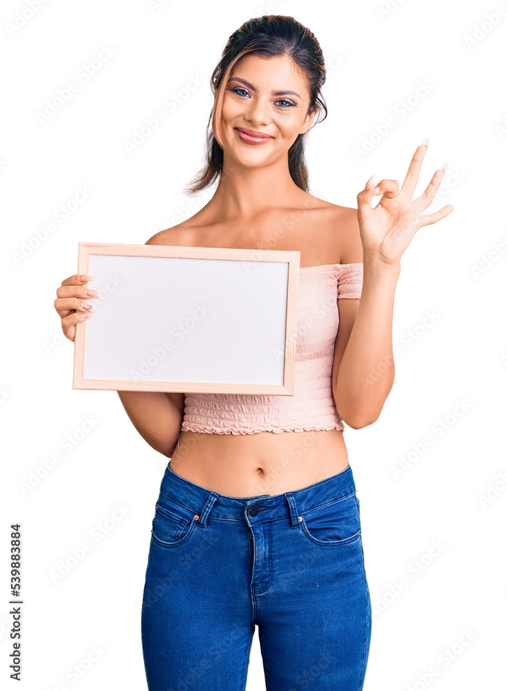 Young beautiful woman holding empty white chalkboard doing ok sign with fingers, smiling friendly gesturing excellent symbol Young beautiful woman holding empty white chalkboard doing ok sign with fingers, smiling friendly gesturing excellent symbol