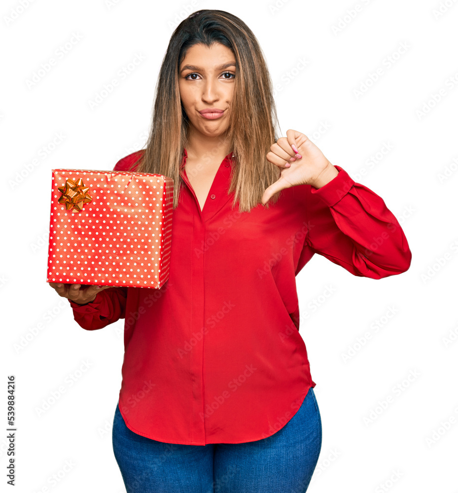 Beautiful brunette woman holding gift with angry face, negative sign showing dislike with thumbs down, rejection concept Beautiful brunette woman holding gift with angry face, negative sign showing dislike with thumbs down, rejection concept