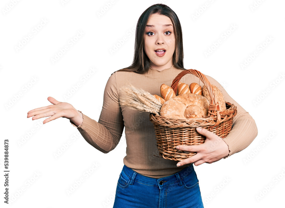 Young hispanic woman holding wicker basket with bread celebrating achievement with happy smile and winner expression with raised hand Young hispanic woman holding wicker basket with bread celebrating achievement with happy smile and winner expression with raised hand