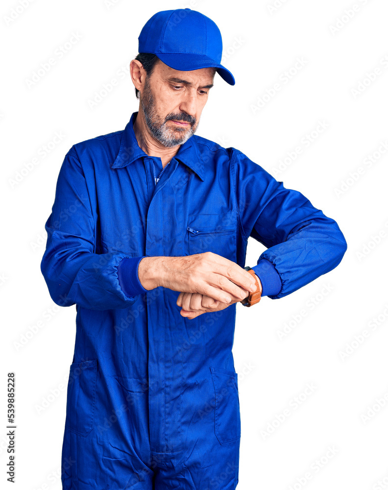 Middle age handsome man wearing mechanic uniform checking the time on wrist watch, relaxed and confident
