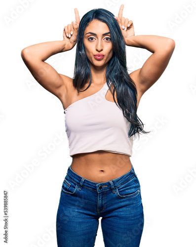 Beautiful hispanic woman wearing casual clothes doing funny gesture with finger over head as bull horns