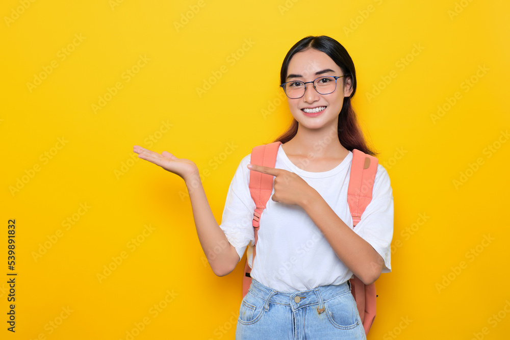 Cheerful pretty young student girl wearing backpack and eyeglasses presenting copy space on palm isolated on yellow background Cheerful pretty young student girl wearing backpack and eyeglasses presenting copy space on palm isolated on yellow background