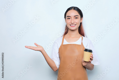 Cheerful pretty young barista girl in apron holding a takeaway cup of coffee, showing copy space on palm isolated on white background