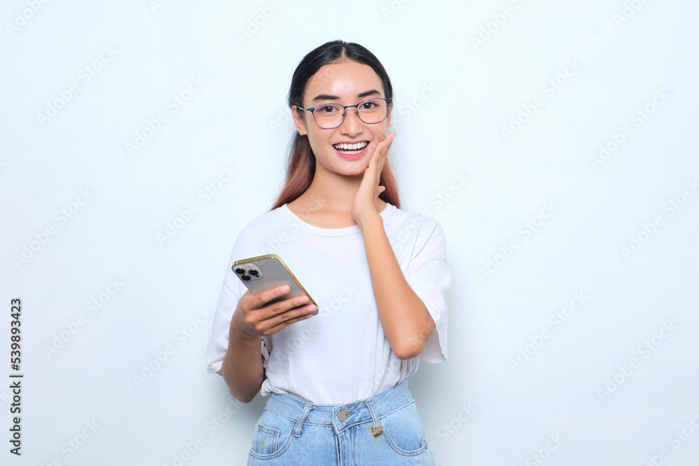 Portrait of smiling young Asian girl in white t-shirt holding mobile phone and touching cheek isolated on white background Portrait of smiling young Asian girl in white t-shirt holding mobile phone and touching cheek isolated on white background