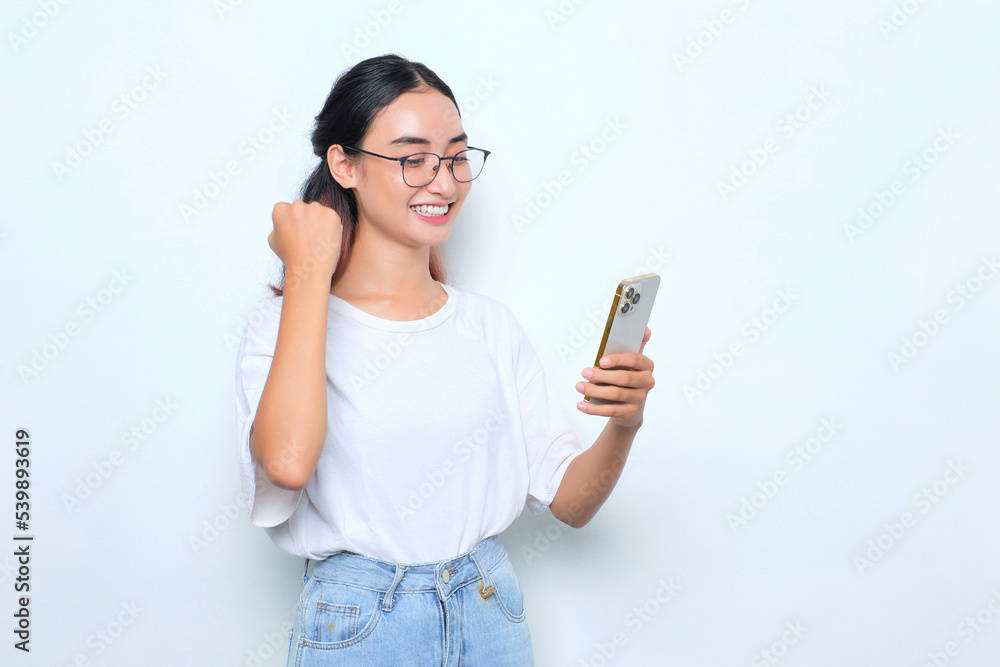 Excited young Asian girl in white t-shirt using smartphone and doing winner gesture isolated on white background