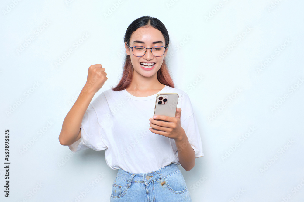 Excited young Asian girl in white t-shirt using smartphone and doing winner gesture isolated on white background Excited young Asian girl in white t-shirt using smartphone and doing winner gesture isolated on white background