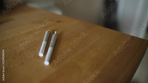 Close up of three marijuana joints on a table. A hand reaches in and picks up one joint and takes it out of frame. photo