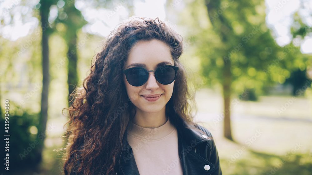 Portrait of attractive young woman in sunglasses and trendy leather jacket looking at camera and smiling standing in city park on summer day. People and sunlight concept.