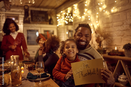 Obraz na płótnie Happy black father and daughter holding placard with 'I'm thankful' while celebrating Thanksgiving with their extended family and looking at camera