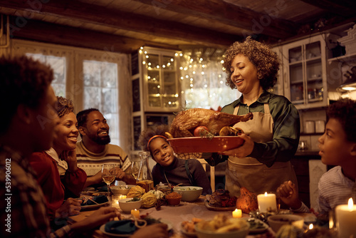 Mature black woman serving traditional Thanksgiving turkey during family meal at dining table.