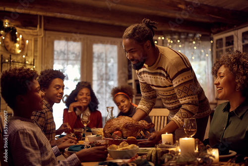 Happy black father serving Thanksgiving turkey to his family at dining table.