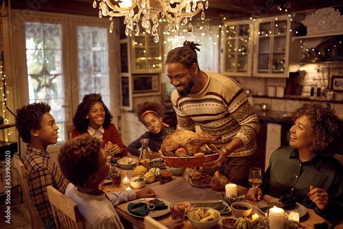 Happy African American man serving stuffed turkey while having dinner with his extended family in dining room on Thanksgiving.