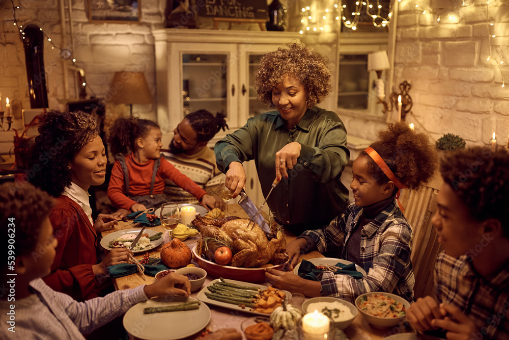 Happy black mature woman carving turkey meat while having Thanksgiving dinner with her family.