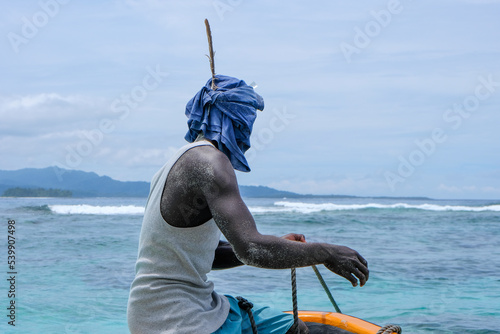 A Melanesian man sitting on the bow of a fishing boat wearing head wrap scarf with a large feather to protect from sun in Bougainville, Papua New Guinea photo