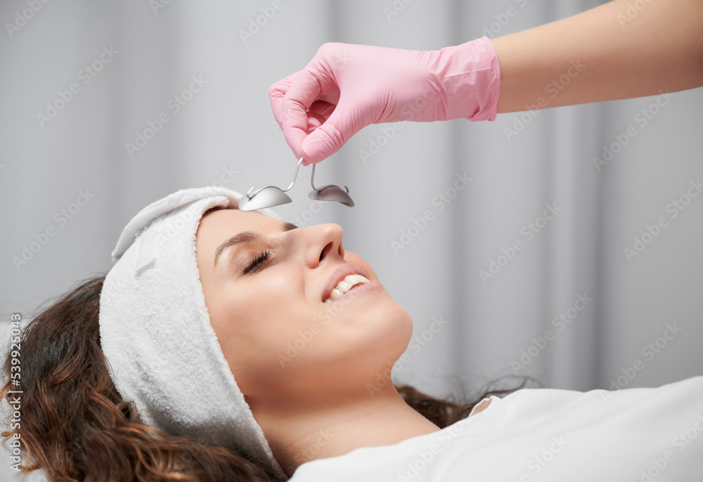 Close up of woman's face who lying with closed eyes at reception at beautician. Cropped view of hand in pink rubber glove keeping metal eye shield from laservision on short distance from face.