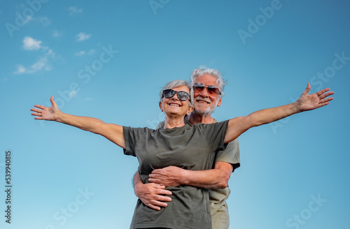 Happy senior couple standing with open arms with blue sky background. Beautiful smiling white-haired seniors enjoying retirement and freedom on an outdoor excursion