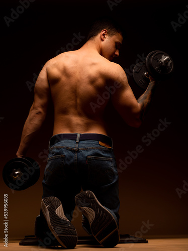 Shirtless young muscular man exercising with dumbbell in gym