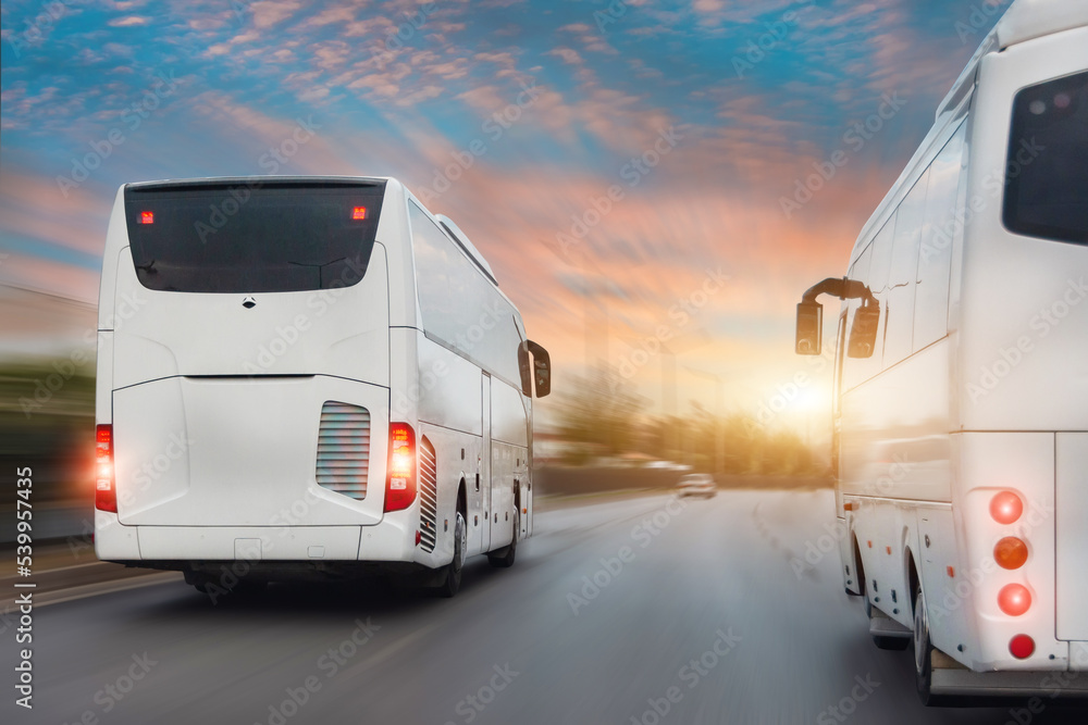 White tourist buses drive along the city highway, overtaking other cars. Traffic in a dense stream of cars with the effect of speed blur in the evening at sunset. White tourist buses drive along the city highway, overtaking other cars. Traffic in a dense stream of cars with the effect of speed blur in the evening at sunset.