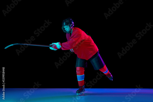 Junior ice hockey player in sports uniform and protective equipment in action over dark background in neon light. Sport, power, challenges, achievement, goals