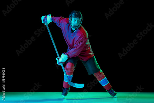 Training of male ice hockey player wearing hockey jersey, uniform and sports helmet in motion isolated over dark background in neon light